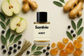 Scentivia Aced It perfume bottle arranged flat on a light cream surface surrounded by halved green apples, whole ginger roots, fresh sage leaves, dark berries, a bundle of dried grass, and pieces of golden amber resin.