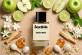 Scentivia First Mist perfume bottle surrounded by whole and halved green apples, lime slices, fresh mint leaves, white jasmine blossoms, pieces of raw wood, golden amber resins, and dried botanicals on a smooth neutral background.