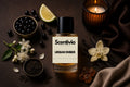 Scentivia Urban Ember perfume bottle surrounded by a small bowl of blackcurrants, a lime wedge, a lit candle in a ribbed amber glass holder, a vanilla orchid flower with vanilla pods, pieces of amber resin, loose black tea leaves in a small bowl, and white jasmine blossoms on a dark brown textured surface.