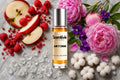 Juicy Crime Roll On bottle surrounded by red apple slices, raspberries, pink peonies, violet flowers, cotton blooms and crystal amber accents on a marble 
background
