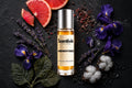 Midnight Dues roll on bottle with grapefruit slices, lavender, purple iris flowers, cotton blooms and peppercorns on dark background